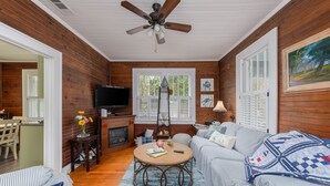 A cozy living room with wooden walls, a ceiling fan, a fireplace, a television, and a dining area with a table and chairs. - Ms. Opal's Cottage - POOL! Pet-Friendly! Ground Level (Tybee Island)