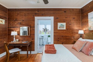 A room with wooden walls, a bed with pink bedding, a desk with a chair, and a window with a view of the outside. - Ms. Opal's Cottage - POOL! Pet-Friendly! Ground Level (Tybee Island)