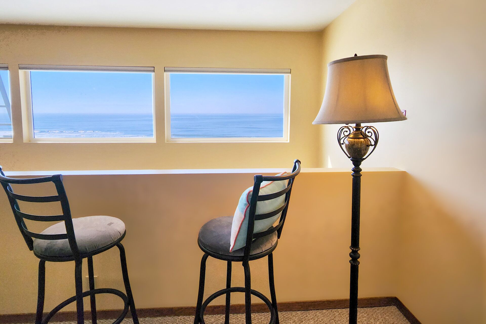 A room with two chairs facing a window overlooking the ocean, a floor lamp, and a beige wall.
