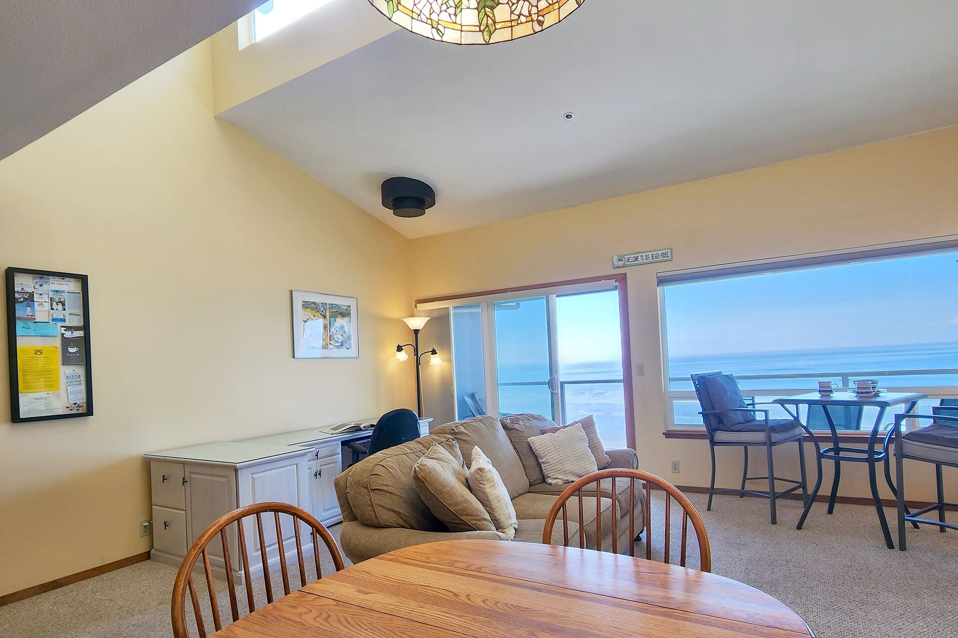 A room with a dining table, chairs, a sofa, a desk, and a large window with a view of the ocean.