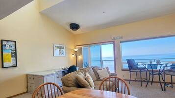 A room with a dining table, chairs, a sofa, a desk, and a large window with a view of the ocean.
