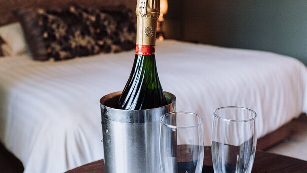 A bed with white linens, a bedside table with a bottle of champagne and two glasses, and a nightstand with a lamp. - Edenmore Golf and Country Club (Craigavon)