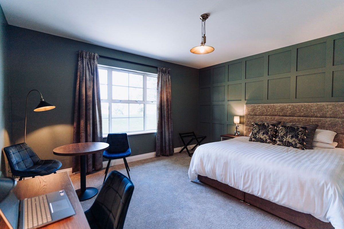 A hotel room with a large bed, a desk with a laptop, a chair, a small table, and a window with curtains.