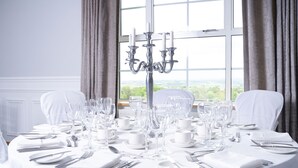 A round dining table set for a formal meal with white tablecloth, silverware, and glassware, positioned near a window with curtains. - Edenmore Golf and Country Club (Craigavon)