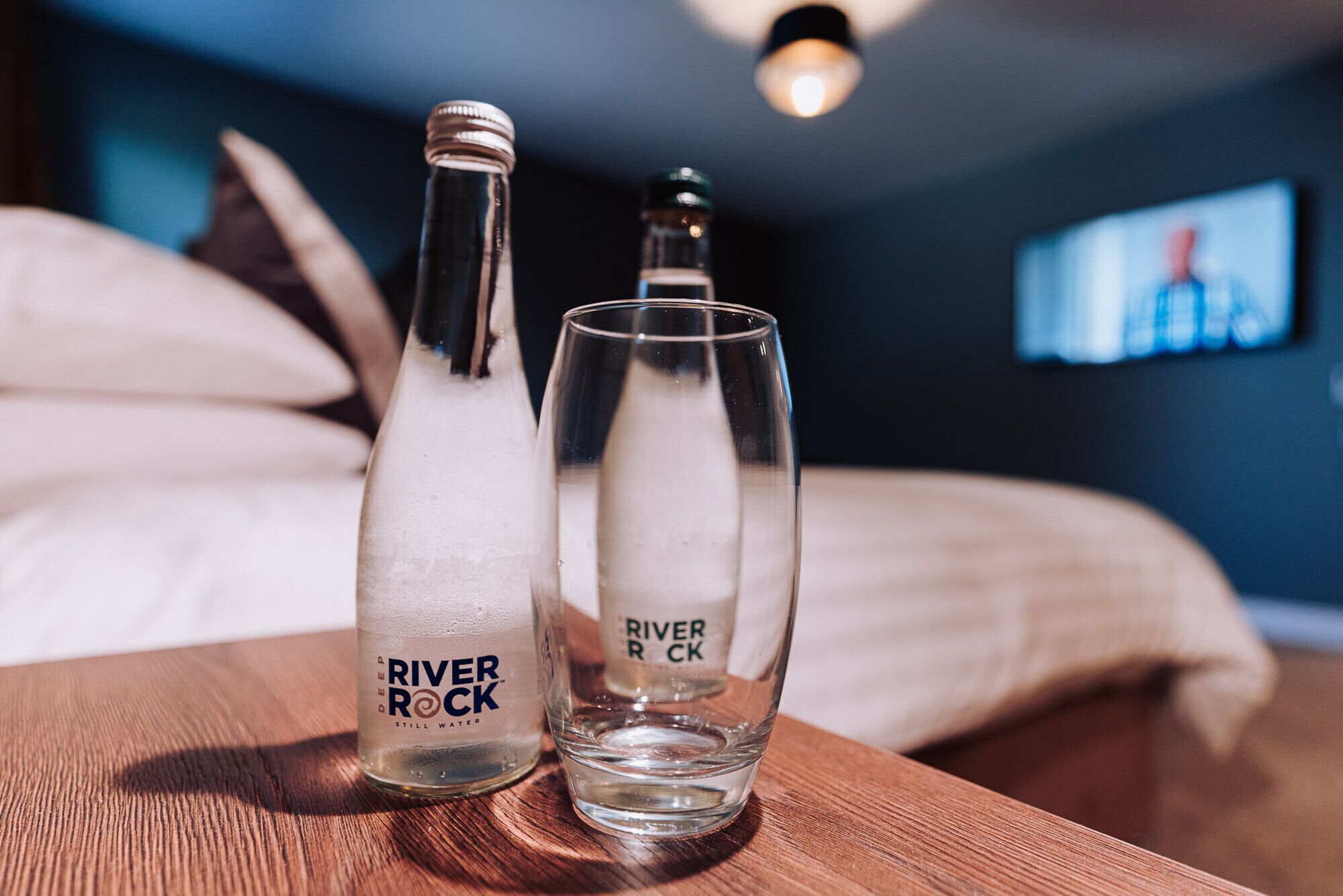 Dos botellas y vasos con la etiqueta 'River Rock' sobre una superficie de madera en una habitación con una cama y un televisor al fondo.