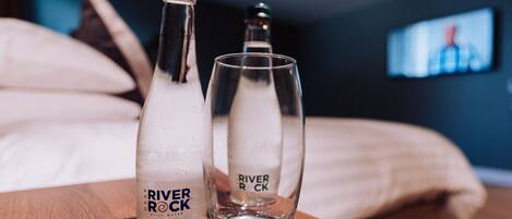 Dos botellas y vasos con la etiqueta 'River Rock' sobre una superficie de madera en una habitación con una cama y un televisor al fondo.