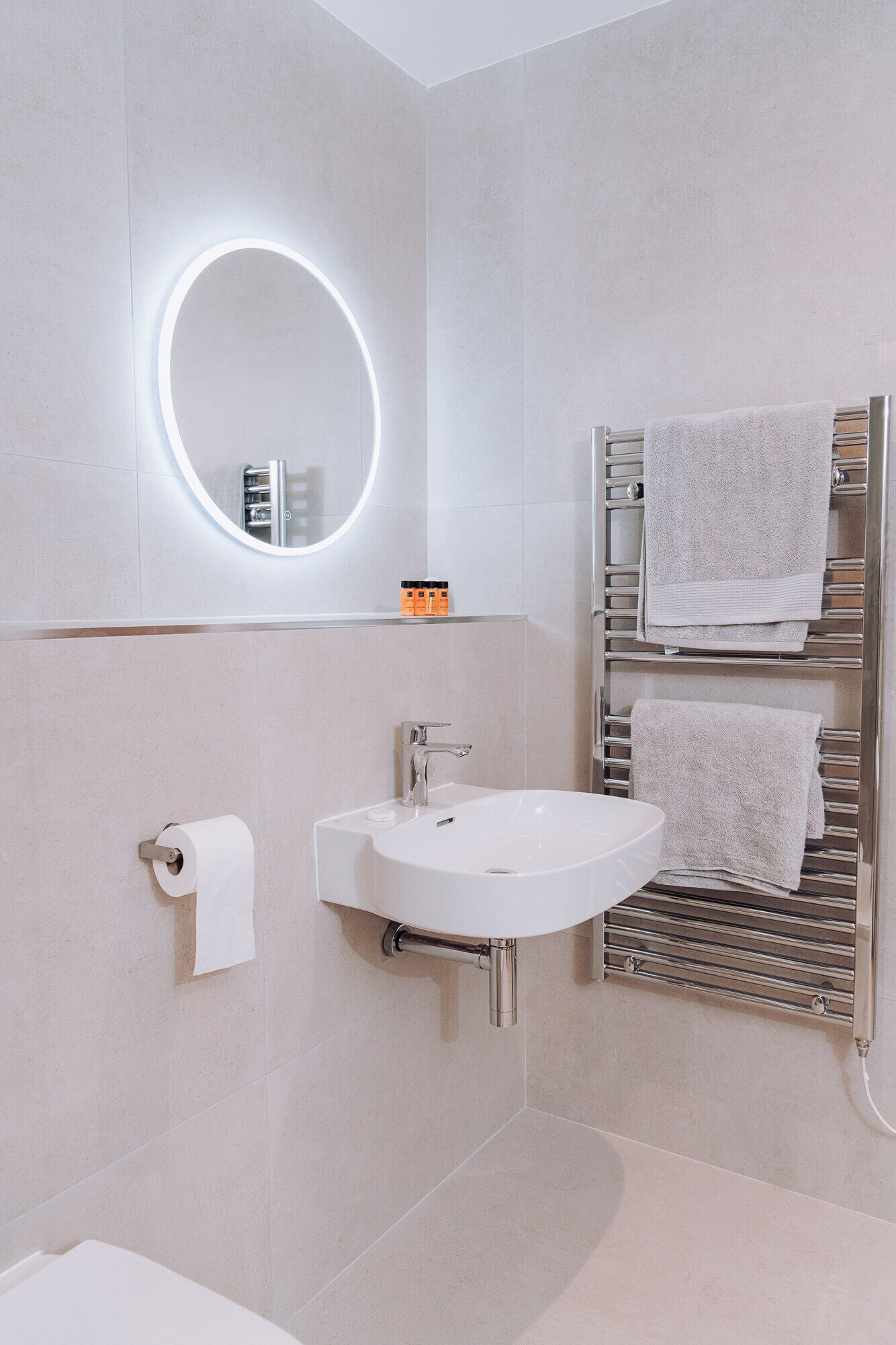 Un baño moderno con un lavamanos blanco, un espejo con luz circular y un perchero con dos toallas.