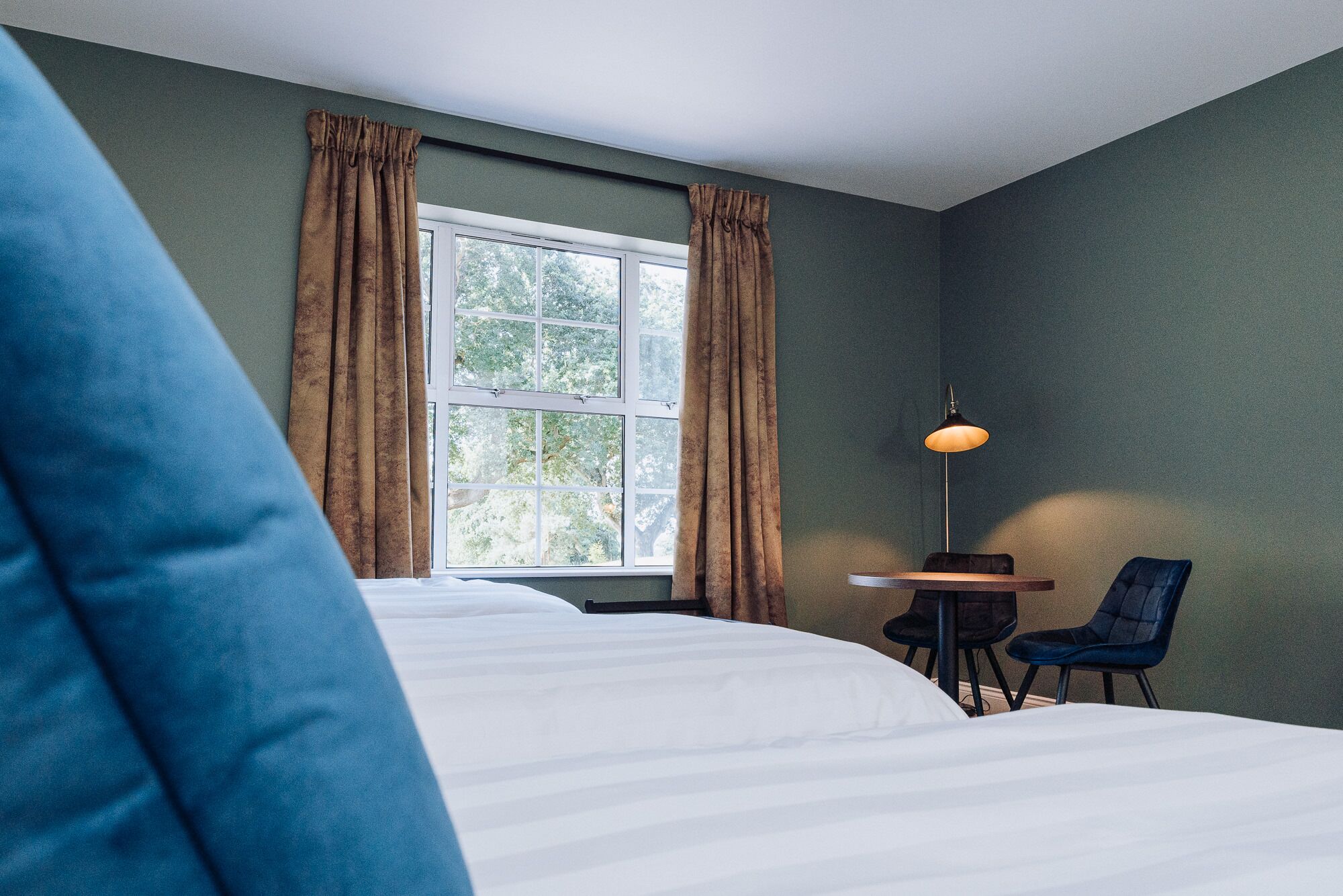 A hotel room with a bed, a desk, two chairs, a window with curtains, and a lamp.
