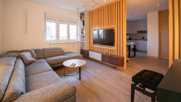 Un soggiorno moderno con un divano componibile, una TV montata su un pannello di legno, un piccolo tavolino e una cucina sullo sfondo.