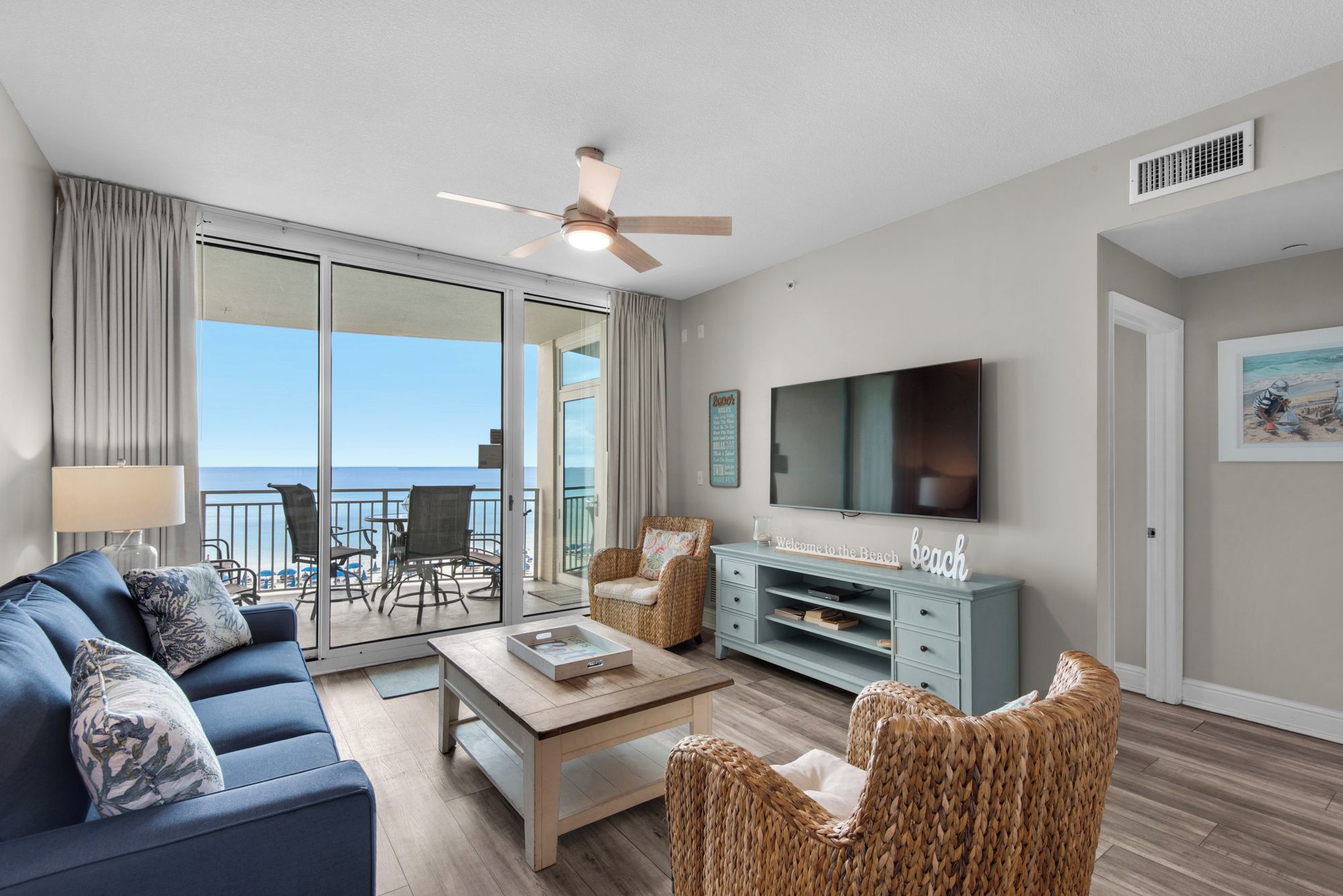 A living room with a sofa, coffee table, wicker chairs, a TV, and a balcony with a view of the ocean.