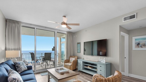 A living room with a sofa, coffee table, wicker chairs, a TV, and a balcony with a view of the ocean.