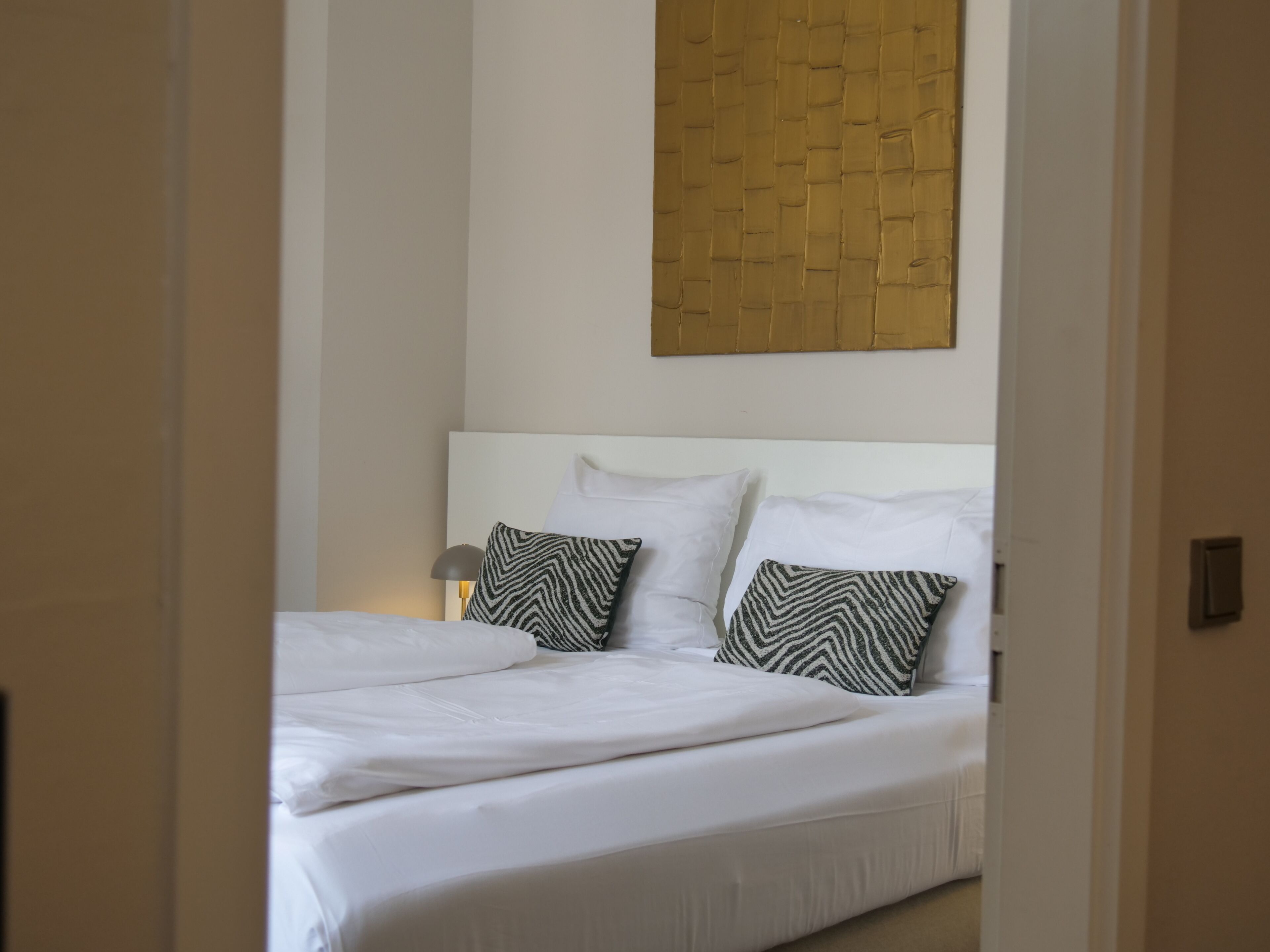 Uma cama bem arrumada com lençóis brancos e duas almofadas estampadas, uma luminária de cabeceira e um painel de madeira na parede acima da cama.