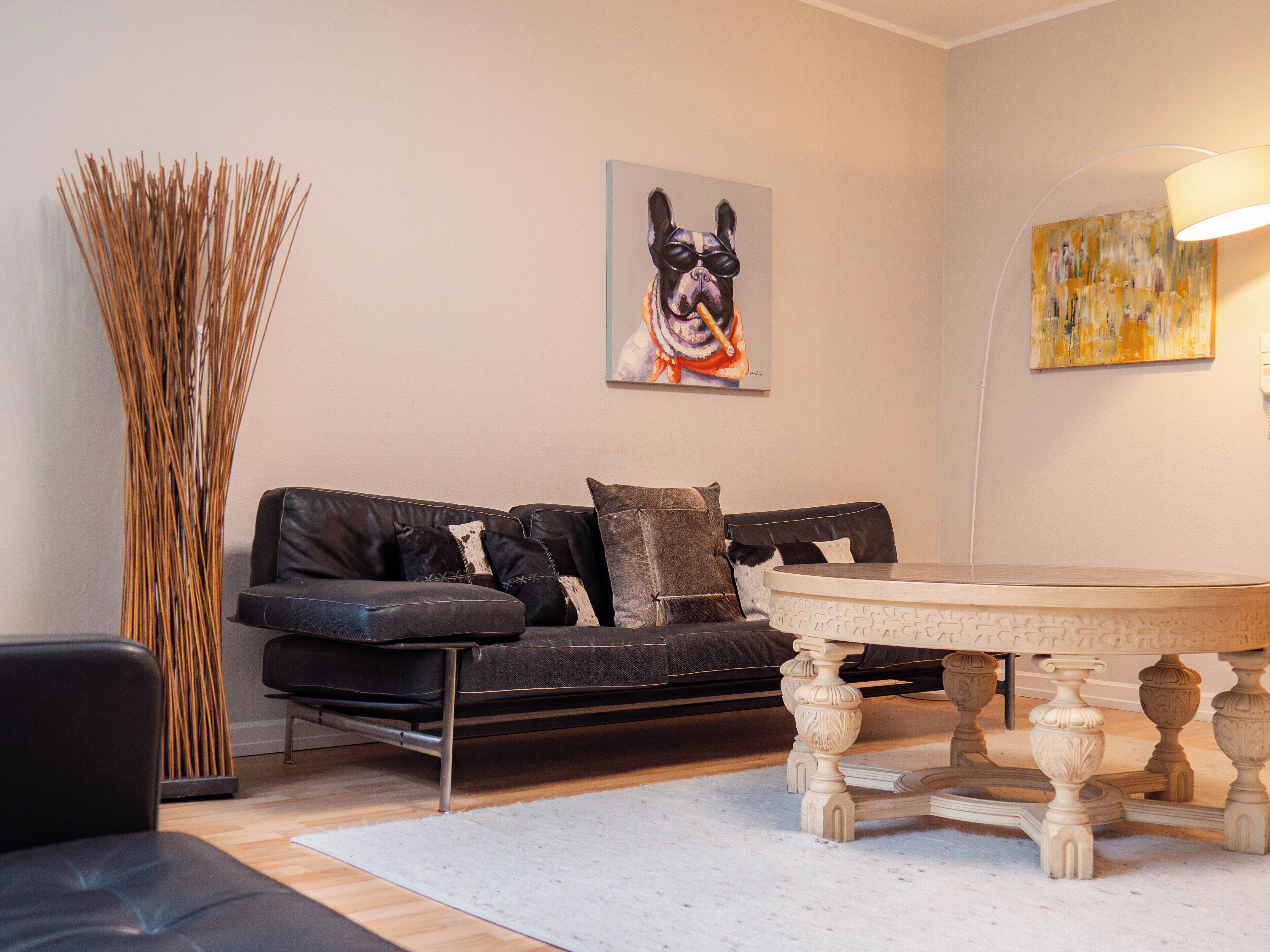 A living room with a black leather sofa, a round wooden coffee table, a tall decorative vase, and a painting of a dog.