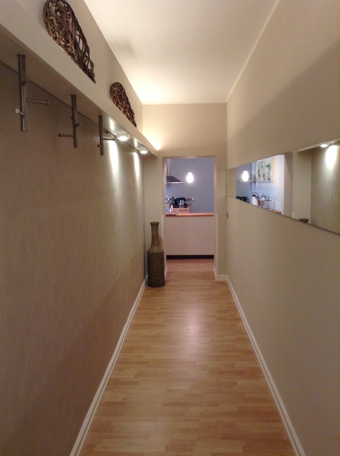 A narrow hallway with wooden flooring, a vase on the floor, and a kitchen visible through an open door.