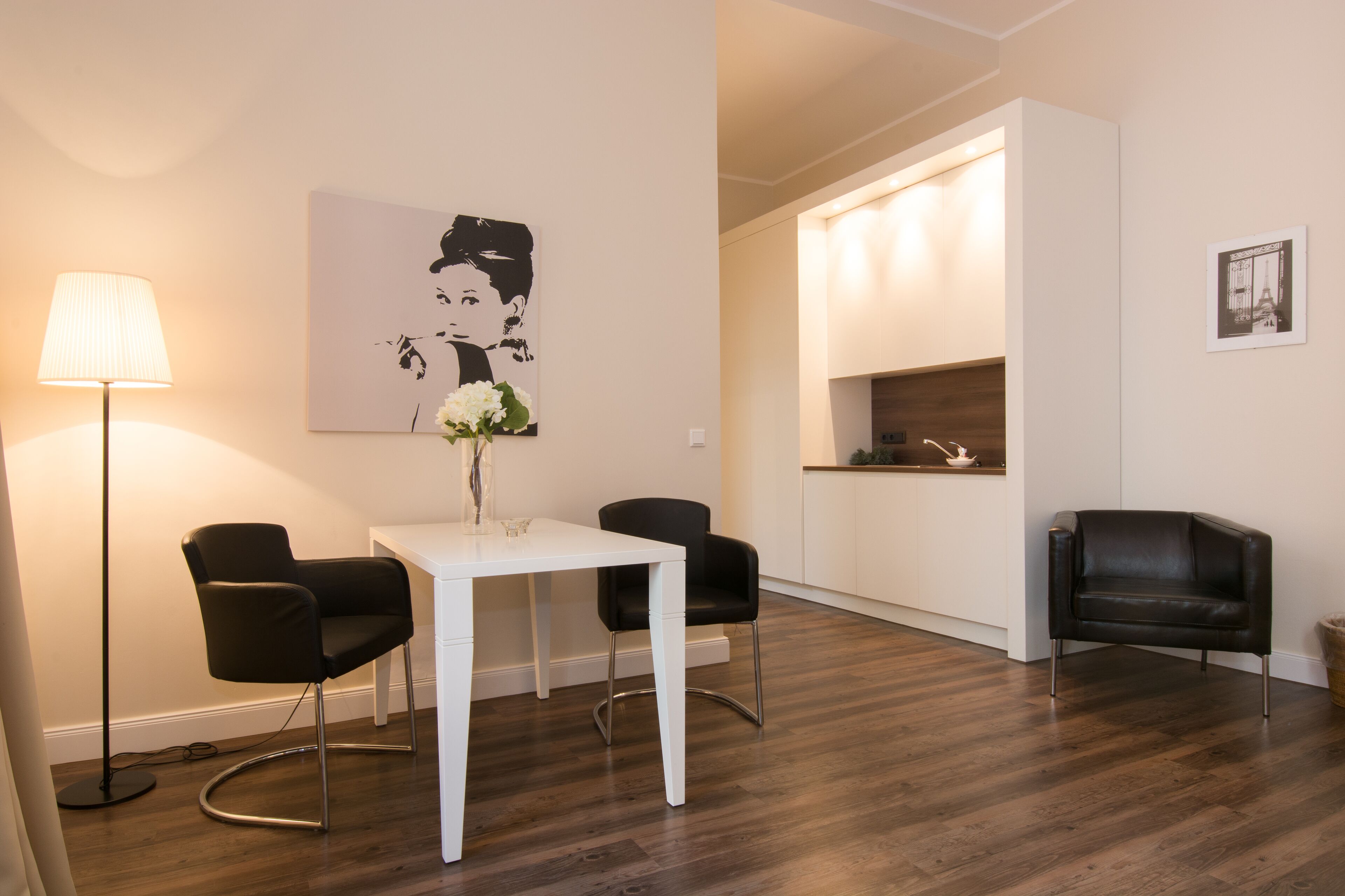 Un salon moderne avec une table blanche, deux chaises noires, un lampadaire et un tableau accroché au mur.