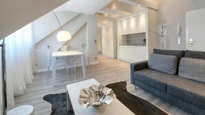 A modern living room with a grey sofa, a white coffee table, and a dining area with a white table and clear chairs. - Living am Carlsplatz (Düsseldorf)