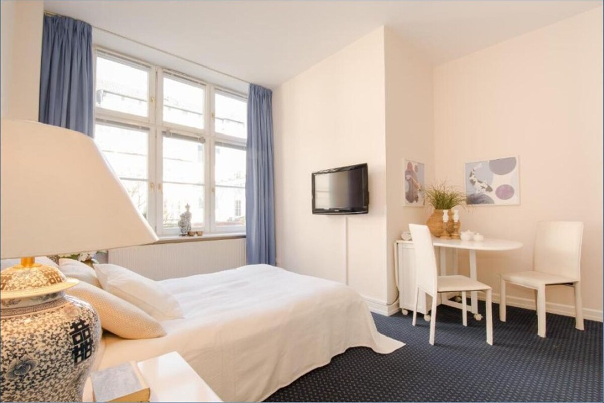 A hotel room with a bed, a desk, two chairs, a TV, and artwork on the wall.
