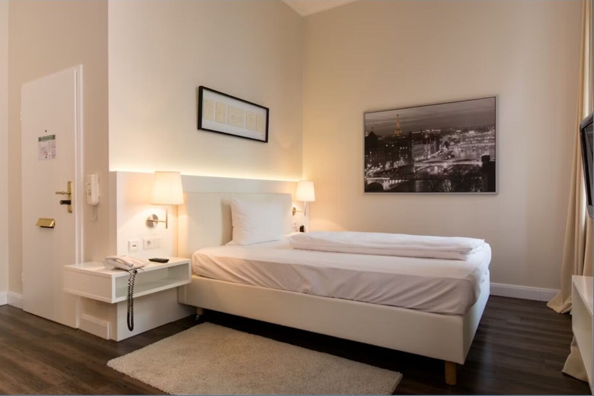 A hotel room with a bed, bedside lamps, a nightstand with a phone, a framed picture on the wall, and a cityscape photo above the bed.