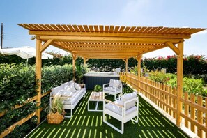 A covered outdoor seating area with a wooden pergola, white outdoor furniture, and a hot tub.