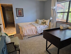 A bedroom with a bed, a desk, a chair, a window with curtains, and a door leading to another room.