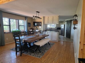 A spacious kitchen with wooden cabinets, a dining table with chairs, and a large window overlooking a forest.