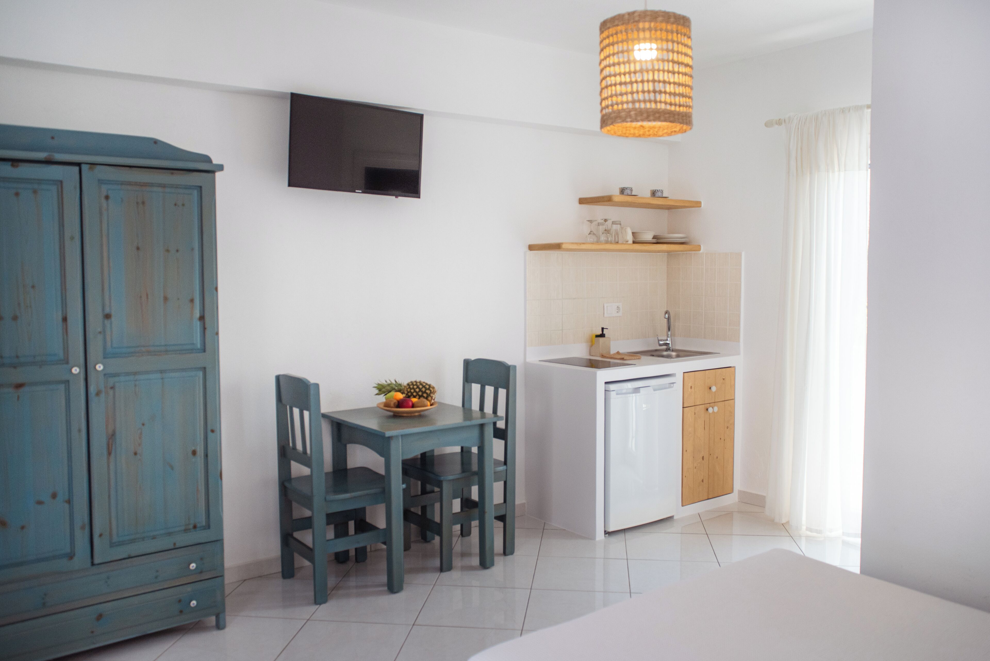 Een compacte keuken met een eethoek, een aan de muur bevestigde televisie en een rieten hanglamp.