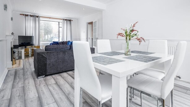 Uma sala de estar moderna com um sofá cinzento, uma televisão e um espaço de jantar com uma mesa e cadeiras brancas.