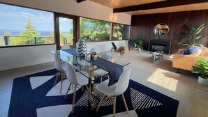 A modern living room with a glass dining table, white chairs, a sofa, and a fireplace. - Pano Water View, Fab Deck, Rec Room, King Bed (Port Angeles)