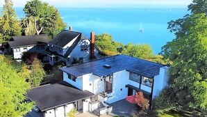 A modern two-story house with a large balcony, surrounded by lush greenery and a view of the sea. - Pano Water View, Fab Deck, Rec Room, King Bed (Port Angeles)