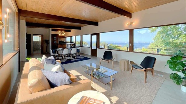 A spacious living area with a large window offering a view of the sea, a modern kitchen, and a seating area with a sofa and chairs. - Pano Water View, Fab Deck, Rec Room, King Bed (Port Angeles)
