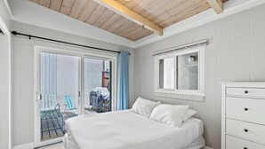 A bedroom with a bed, a dresser, a window, and a sliding glass door leading to an outdoor area with chairs. - Heart of Capitola Vacation Rental (Capitola)
