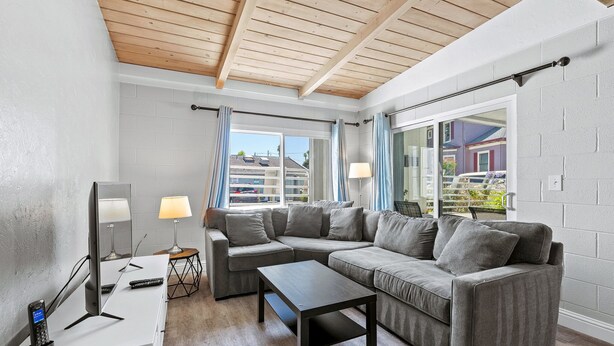 A modern living room with a sectional sofa, a coffee table, and a desk with a computer and phone. There are windows with curtains, a lamp, and a view of buildings outside. - Heart of Capitola Vacation Rental (Capitola)