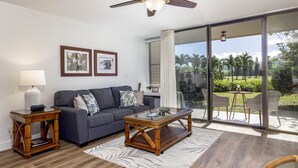 A living room with a sofa, coffee table, and side tables, a ceiling fan, and a large sliding glass door leading to an outdoor patio. - Turtle Bay Golf Course View. Updated 1-bed w/*AC, covered lanai, end unit (Kahuku)
