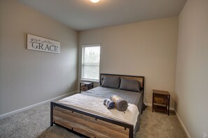 A bedroom with a bed, a bedside table, a nightstand, a window with blinds, and a wall sign. - Romantic 3-BR Townhouse With Backyard ~ Double Story - Pet Friendly! (Rosenberg)