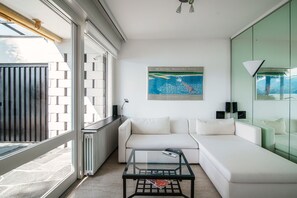 A modern living room with a glass coffee table, a white sofa, and a large window with a view of the city.