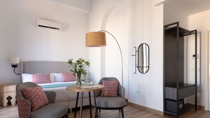 Ett modernt hotellrum med en säng, två fåtöljer, ett litet runt bord, en golvlampa, en spegel och en inbyggd TV.
