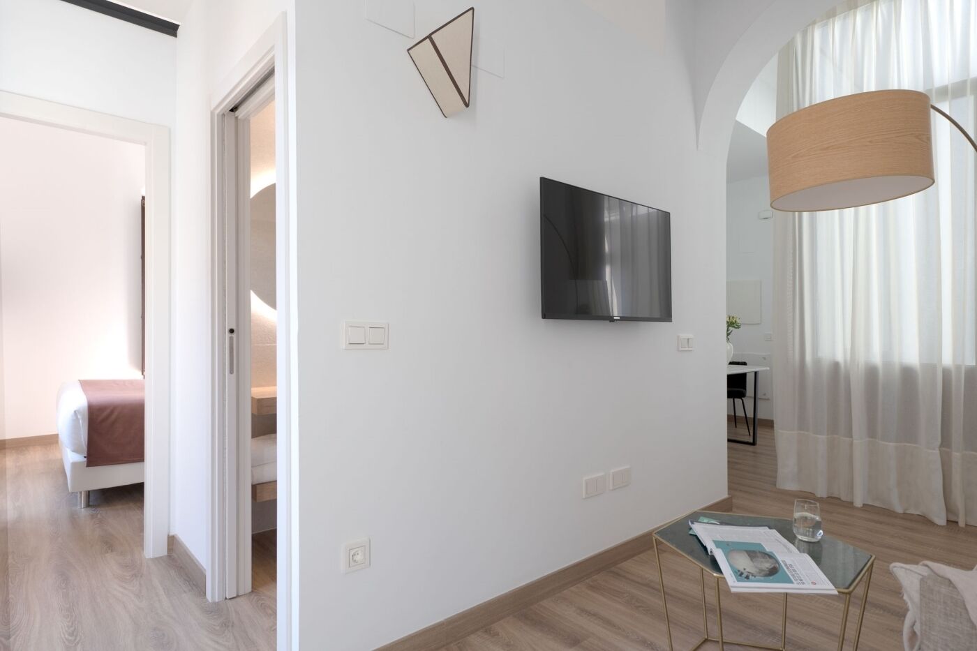 Quarto moderno e bem iluminado, com televisão de tela plana na parede, mesa de vidro com uma revista e piso de madeira.