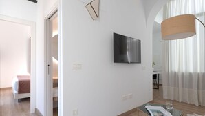 Une pièce moderne et bien éclairée, dotée d’un téléviseur à écran plat fixé au mur, d’une table en verre avec un magazine et d’un plancher en bois.