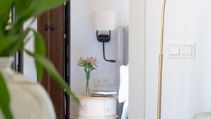 Een kamer met een houten deur, een witte muur met een wandlamp, een klein wit tafeltje met een bloevas en een stoel.