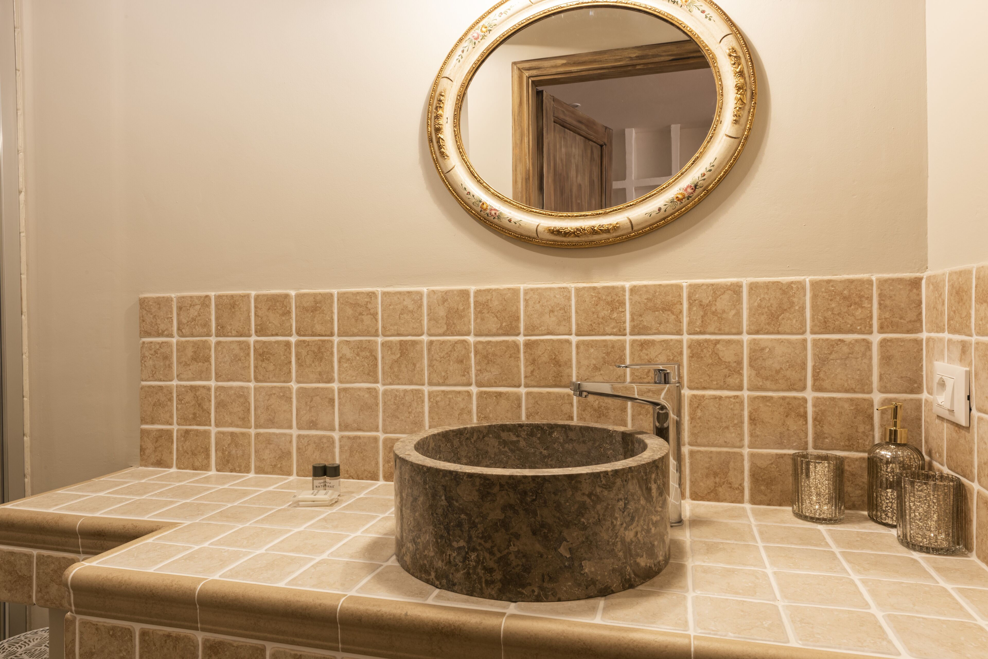 A bathroom with a round sink, a mirror with a gold frame, and a tiled backsplash.