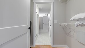 A narrow hallway with a washing machine and a drying rack, leading to a room with a door.