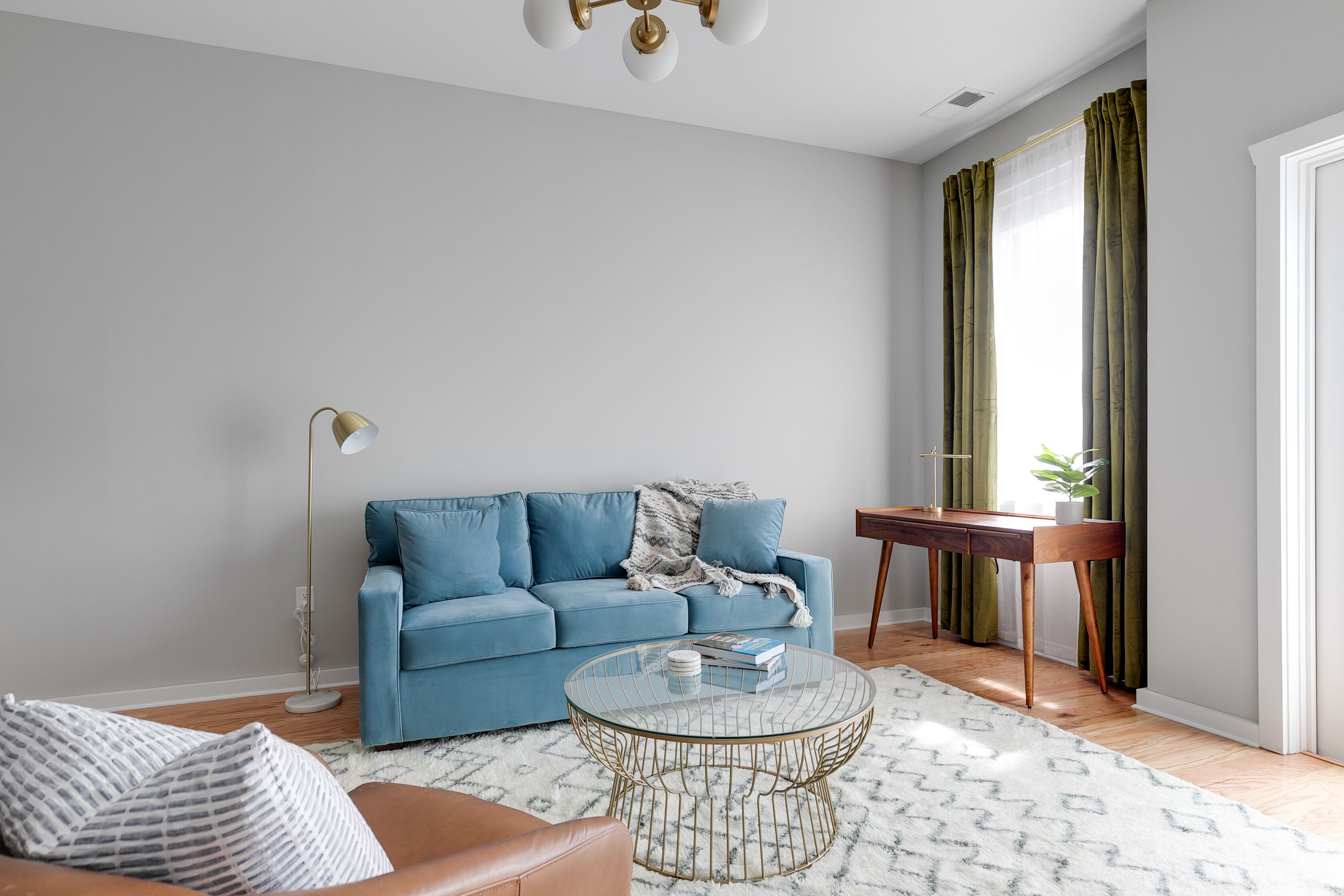 Una sala de estar con un sofá azul, una mesa de centro de vidrio, un sillón de cuero marrón, una lámpara de pie y una mesita de noche con una planta.