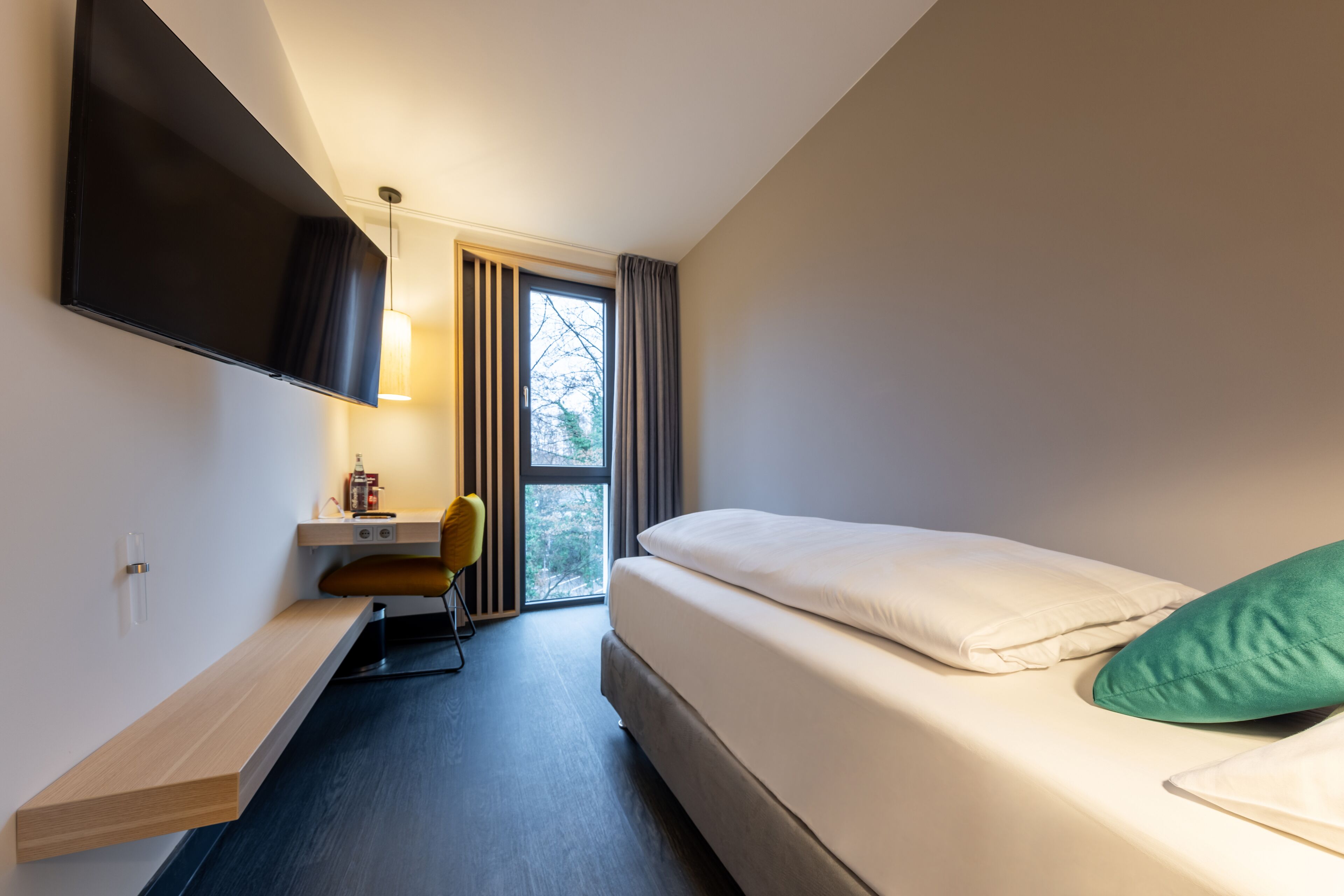 Ein modernes Hotelzimmer mit Bett, Schreibtisch, Stuhl, Fernseher und einem Fenster mit Vorhängen.