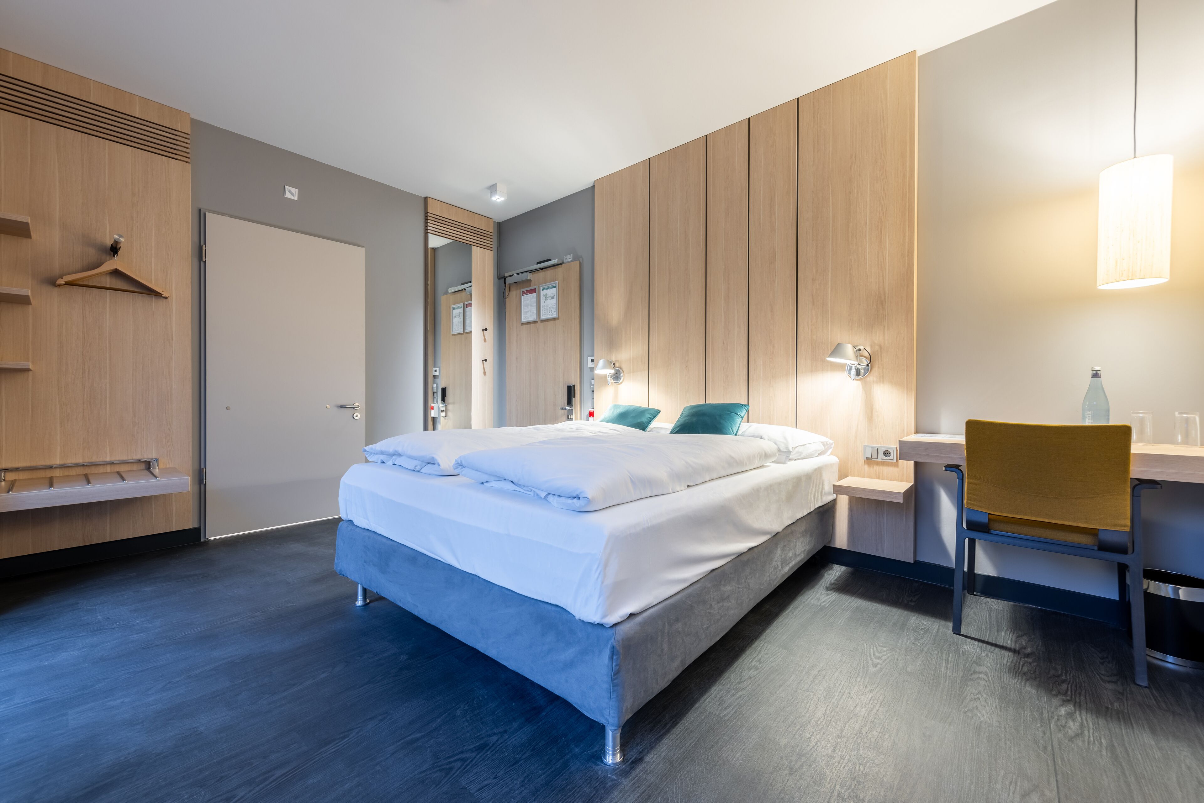 A modern hotel room with a bed, a desk, a chair, a wardrobe, and a mirror.