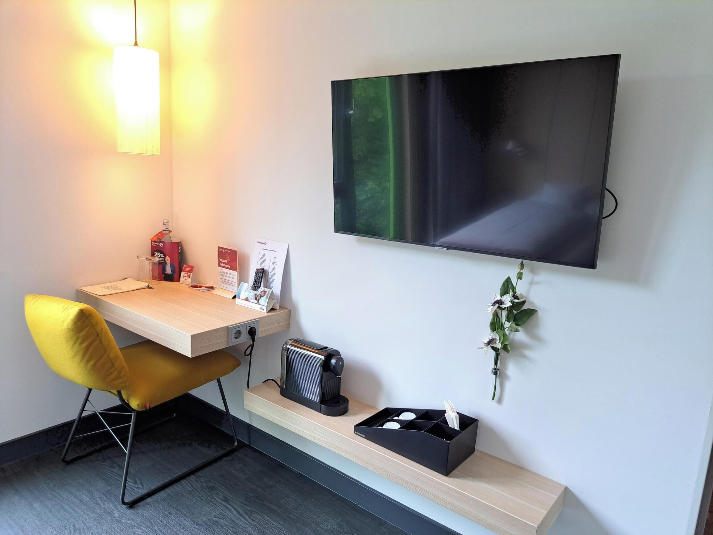 Ein modernes Hotelzimmer mit Schreibtisch, flachbildfernseher an der Wand, einem kleinen Heizkörper und einer Ablage mit Blumen in einer Vase.