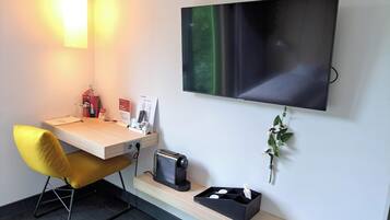 Ein modernes Hotelzimmer mit Schreibtisch, flachbildfernseher an der Wand, einem kleinen Heizkörper und einer Ablage mit Blumen in einer Vase.