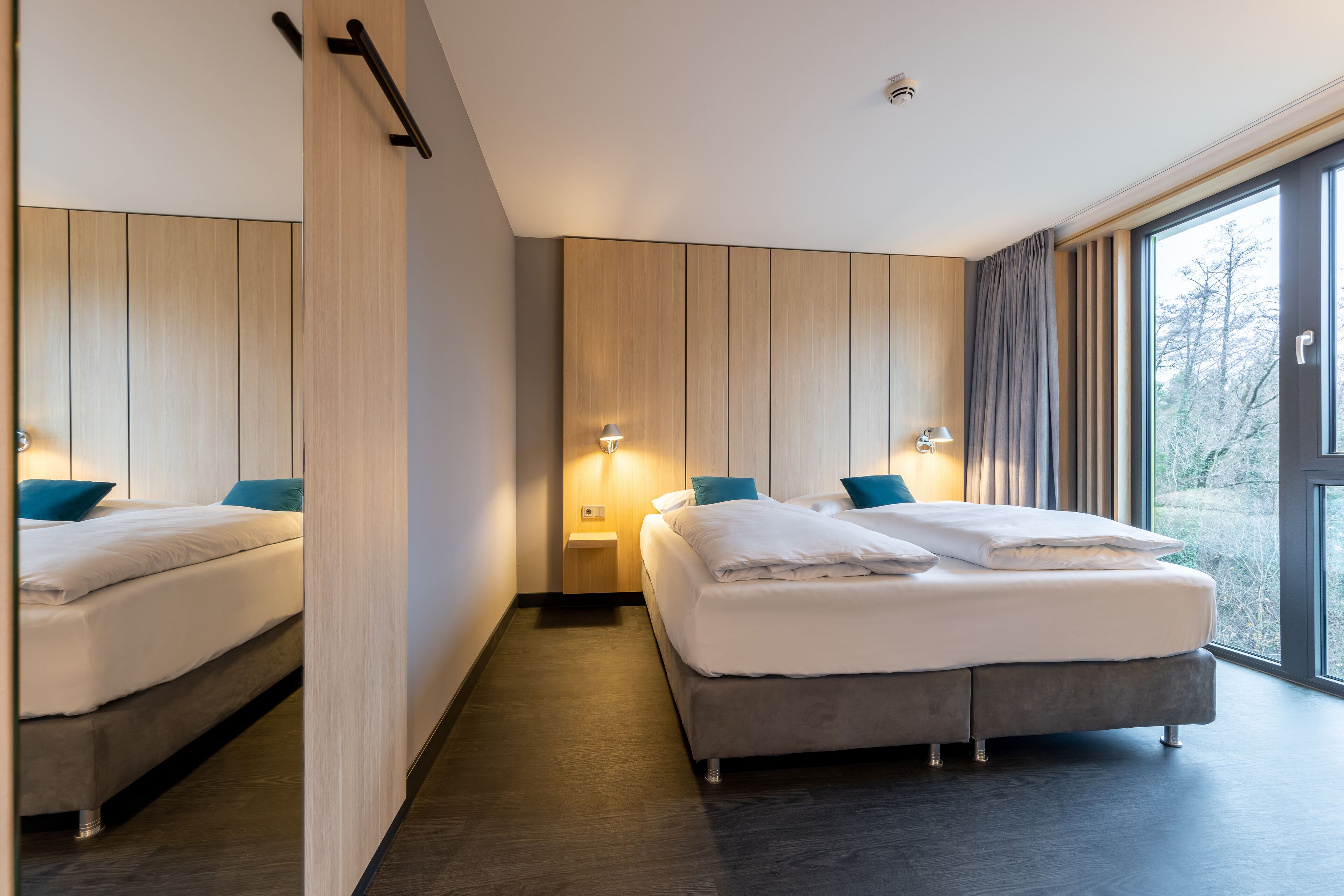 Una habitación de hotel moderna con dos camas, paneles de madera en las paredes y grandes ventanales con vistas al exterior.