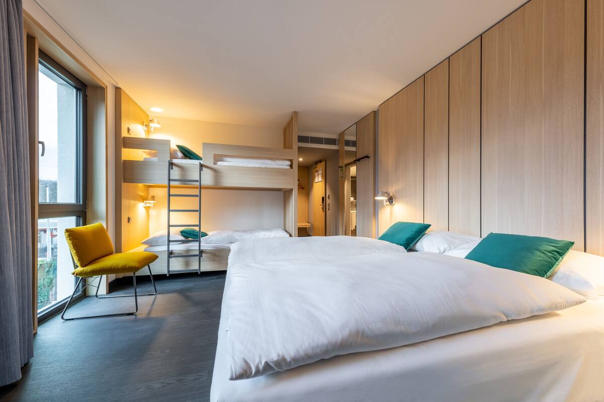 A hotel room with a large bed, a yellow chair, a ladder, and a view of the outdoors.