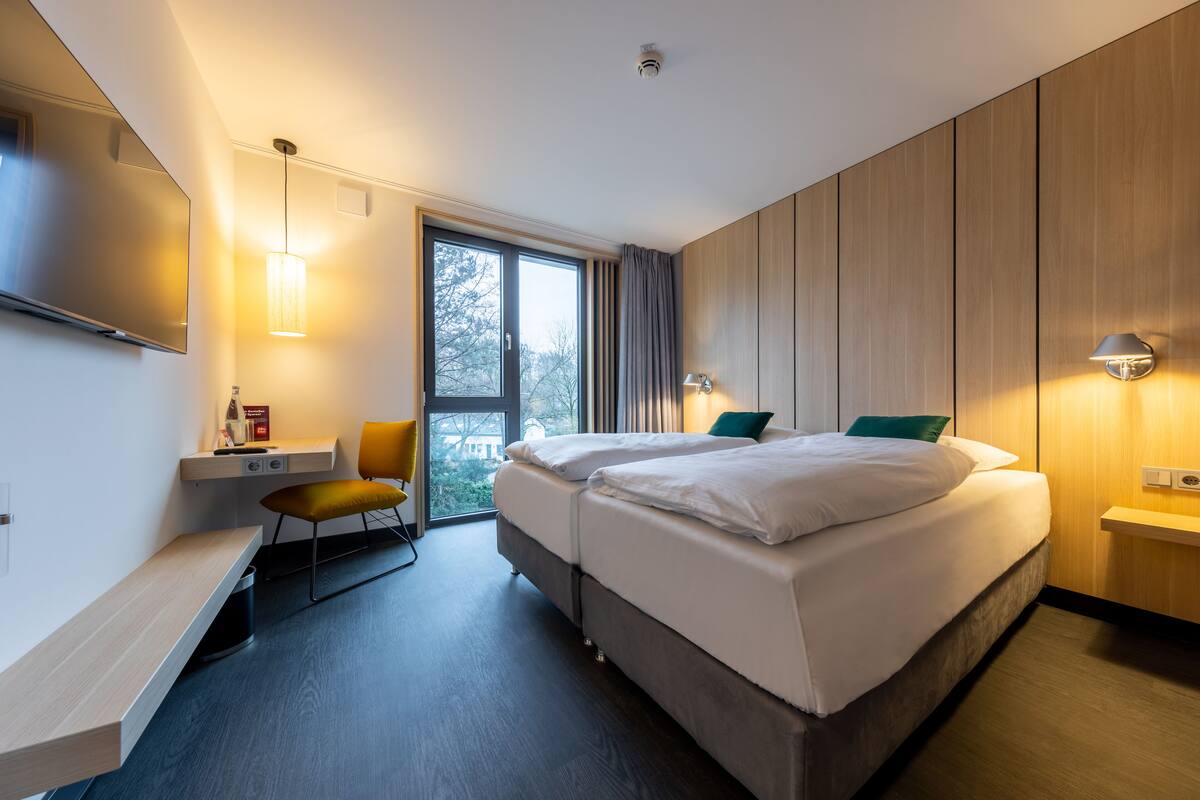 A modern hotel room with a large bed, a desk, a chair, and a window with a view.
