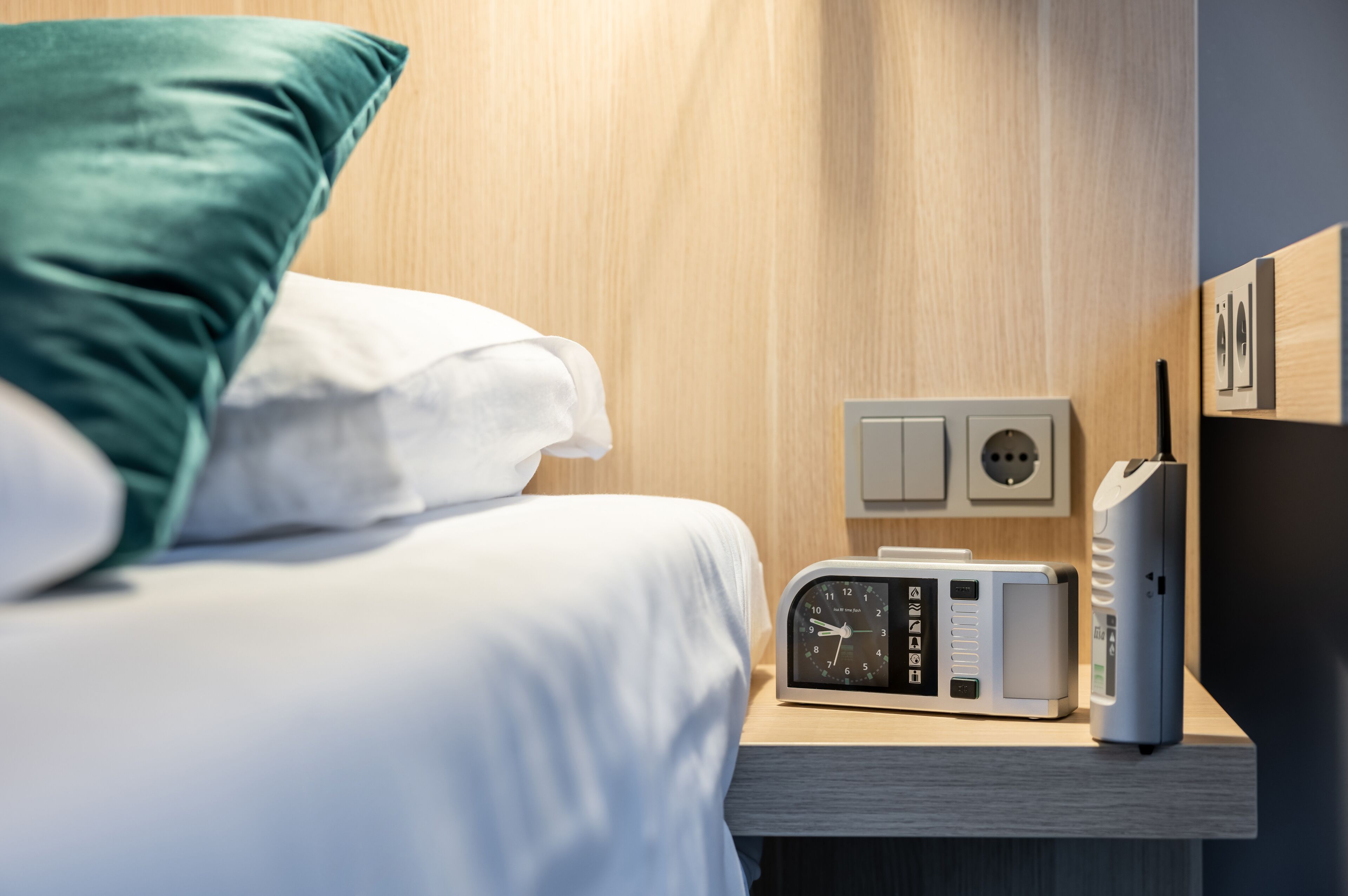 Ein ordentlich bezogenes Bett mit einem grünen Kopfkissen, ein Nachttisch mit einer Uhr, einem Mehrfachsteckdosenadapter und einem Ladegerät.